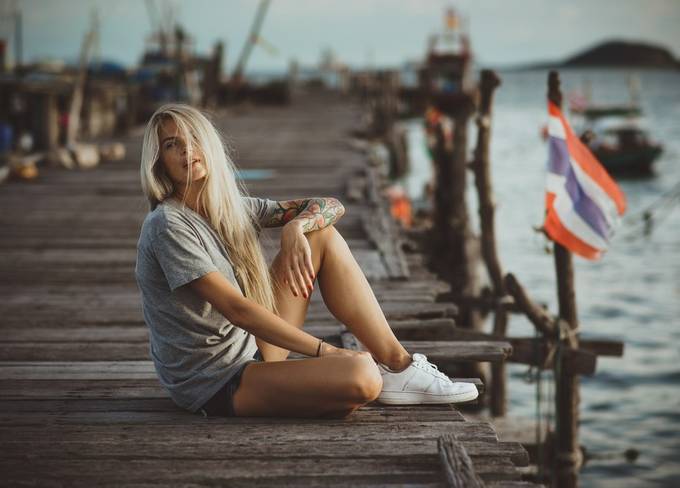 Hipster girl sitting on wooden pier in the rays of the setting sun. A vacation in Thailand. Hands with tattoos. by cinematheart - The Lifestyle Project